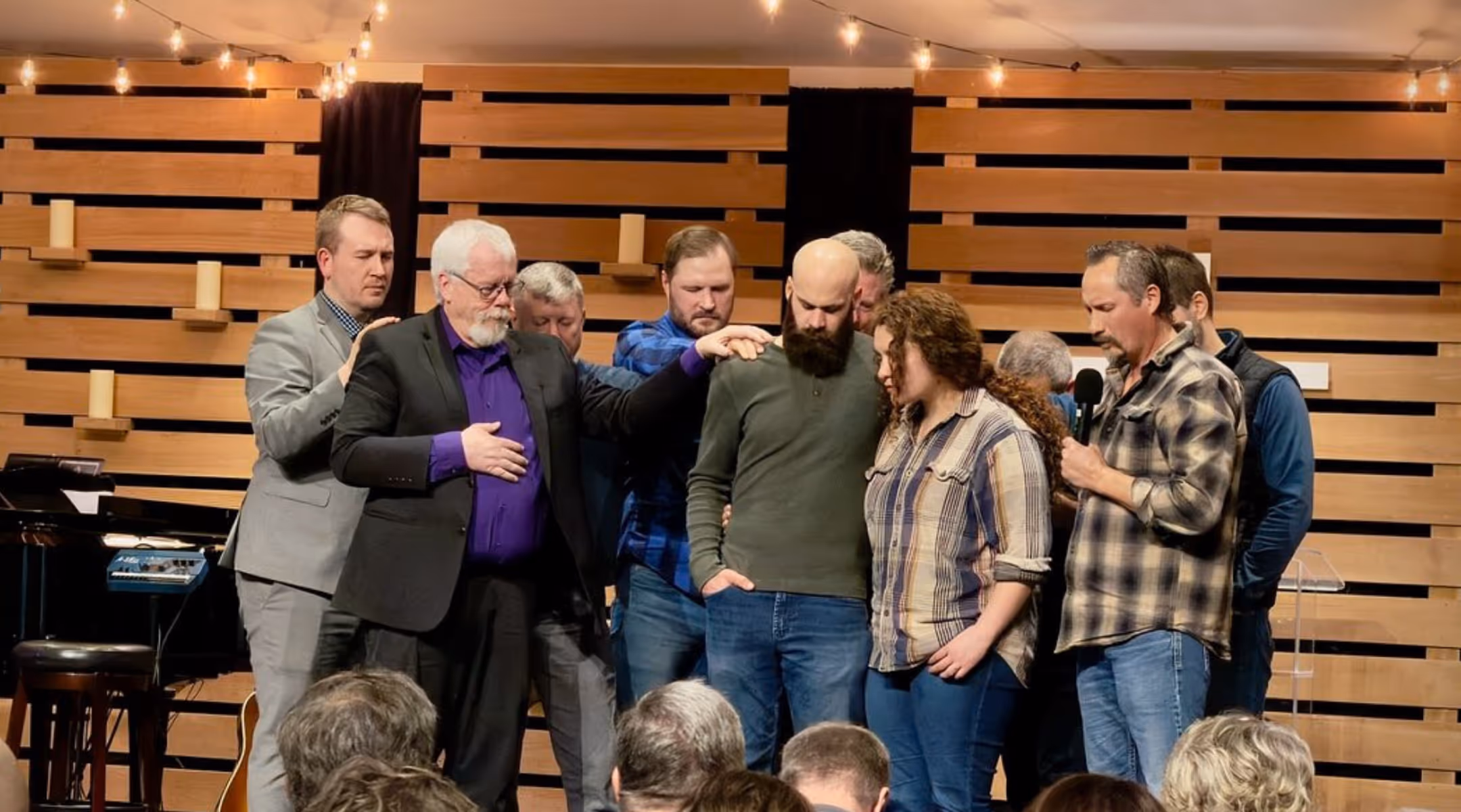 Group of people standing on stage while some lay hands on two individuals in a supportive or prayer gesture.