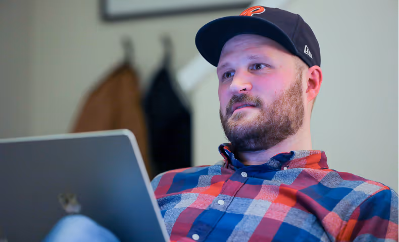 Bearded man wearing a baseball cap and plaid shirt, looking at a laptop screen.