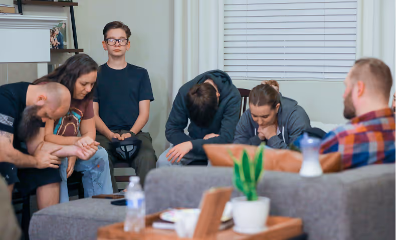 A group of people sitting closely together in a living room with heads bowed and eyes closed, appearing to pray or reflect.
