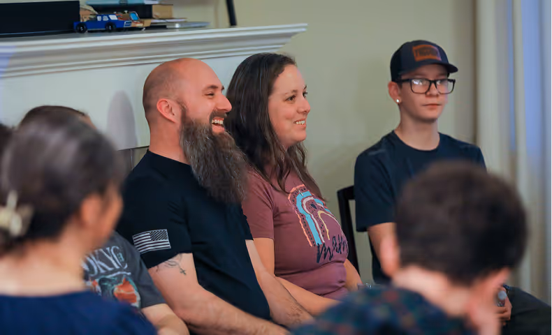 Group of people sitting indoors, with a bearded man and a woman smiling in the center, and two others partially visible.