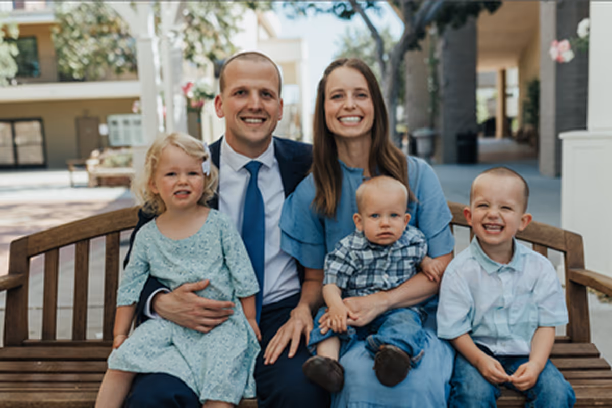 Smiling family of five sitting on a wooden bench outdoors, with father holding daughter, mother holding baby, and young boy seated next to them.