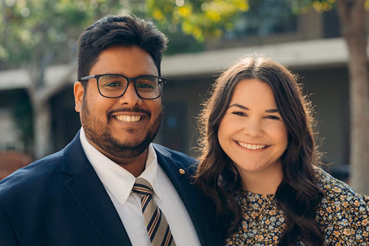 Smiling man in glasses and suit with woman in floral dress outdoors with blurred trees in background.