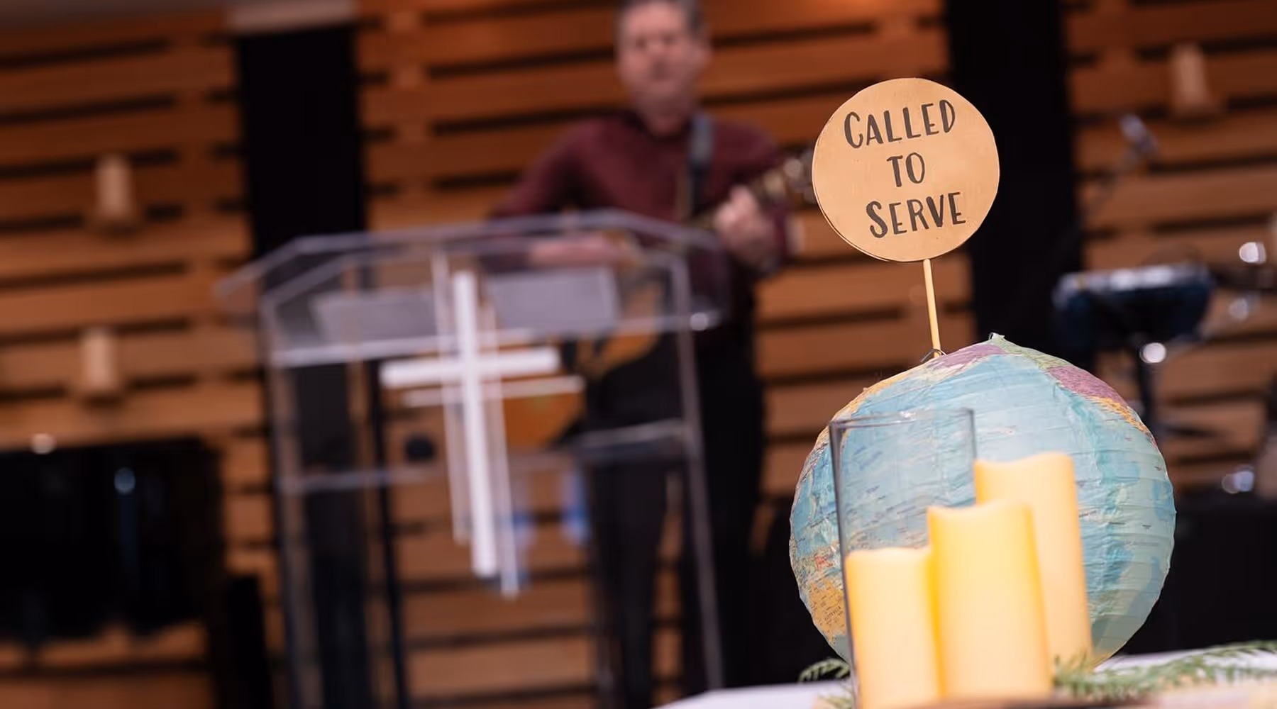 Interior of a church with a globe featuring a sign that says 'Called to Serve' and candles in the foreground, with a person playing guitar blurred in the background.
