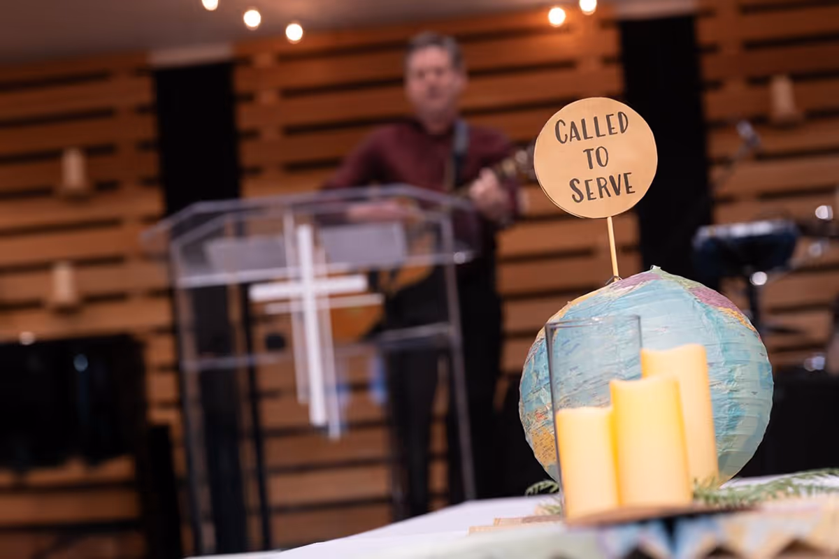 Decorative globe with a sign saying 'CALLED TO SERVE' and candles on a table, with a blurred man playing guitar behind a podium with a cross in a church setting.