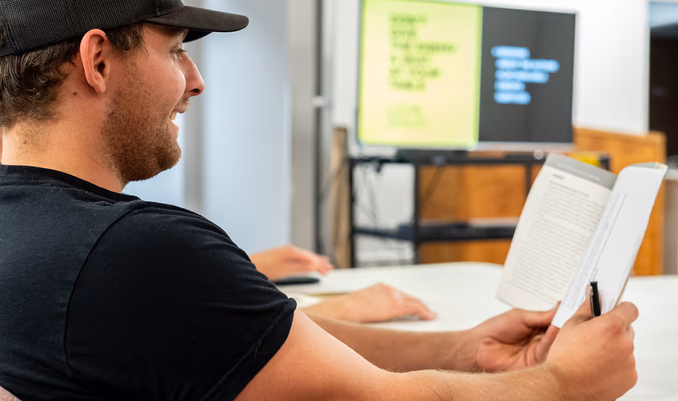 Young man wearing a black cap and t-shirt reading a book in a classroom with a blurred lesson on a screen in the background.