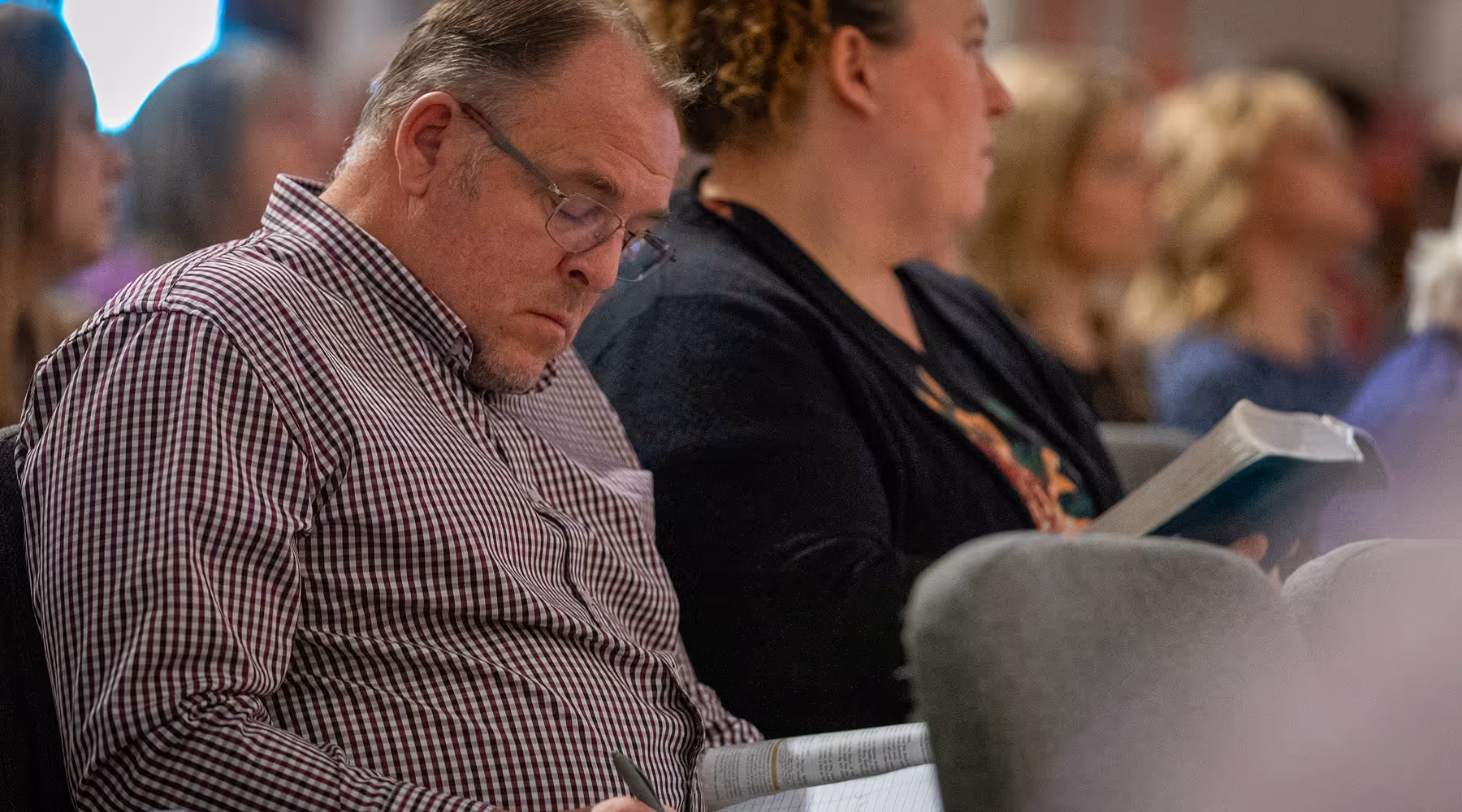 Man in glasses and checkered shirt writing in a notebook while sitting next to a woman holding an open book in a crowded room.