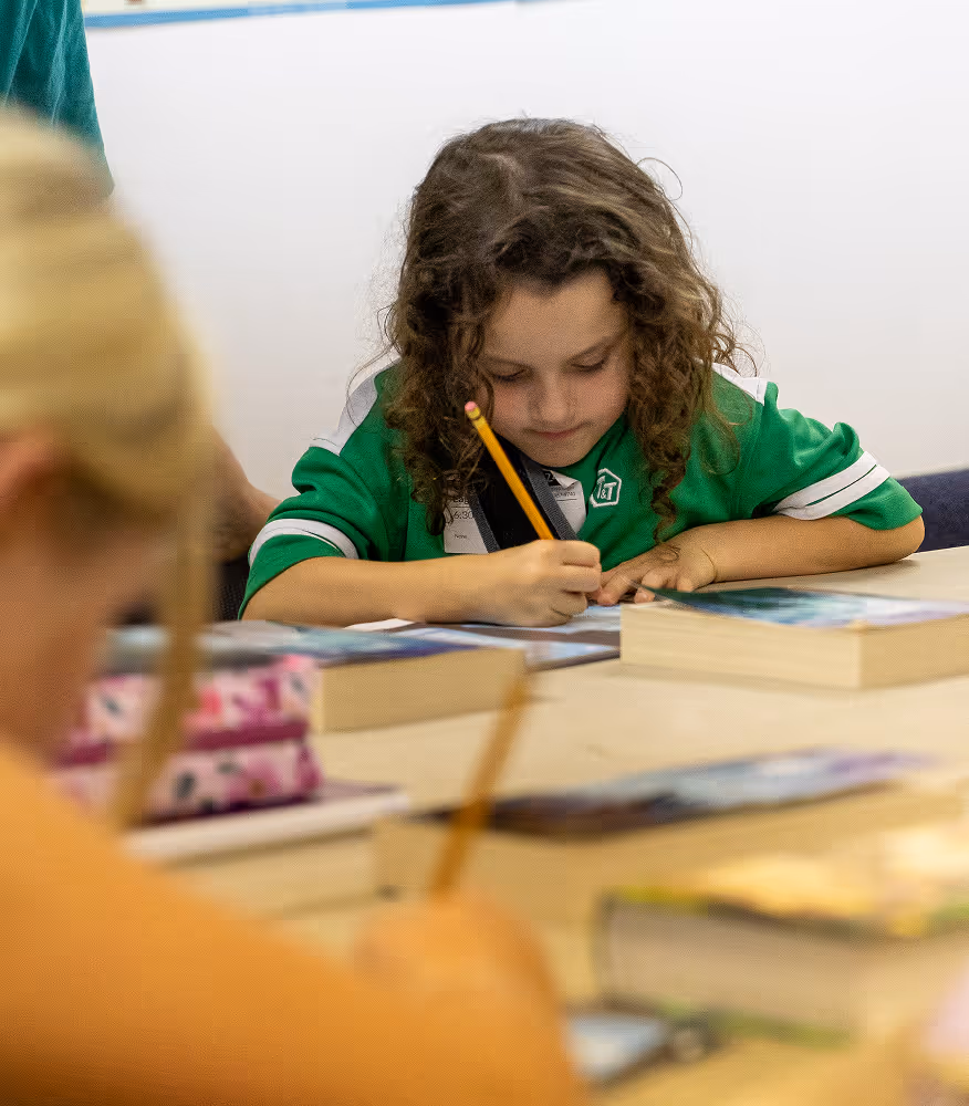 Young girl with curly hair writing with a pencil at a table with books, focused on her work.