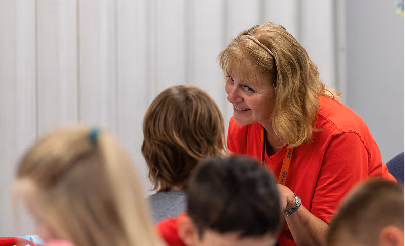 Female teacher in a red shirt smiling and talking closely with a student in a classroom setting.