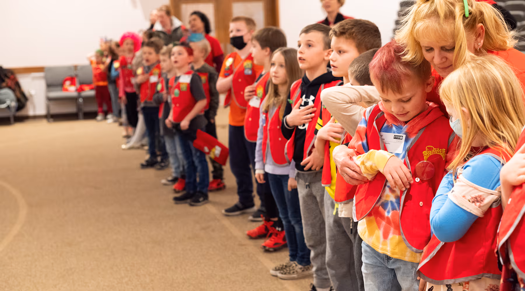 A group of children wearing red vests standing in a line indoors, some with hands over their hearts, and a woman hugging two children in the foreground.