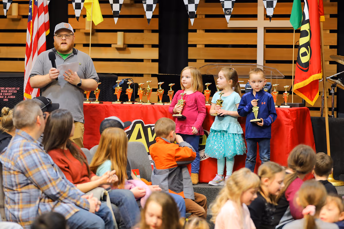 Three young children holding trophies on stage during an awards ceremony, with an adult speaker holding a microphone beside them and an audience watching.