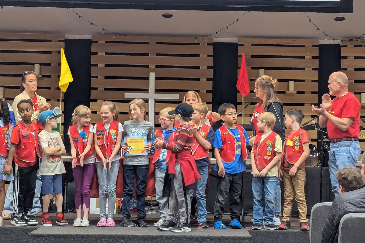 Group of children wearing red vests standing on a stage with adults applauding, with a cross and flags in the background.