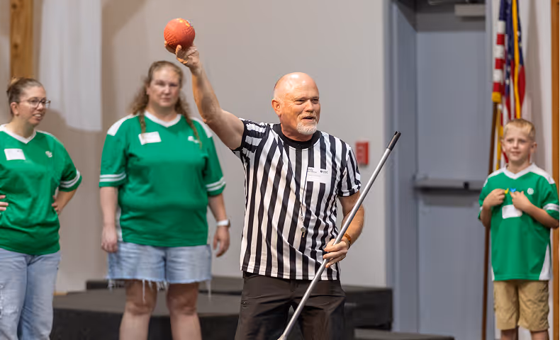 Referee holding a red ball and a pole, with three people in green shirts watching in the background.