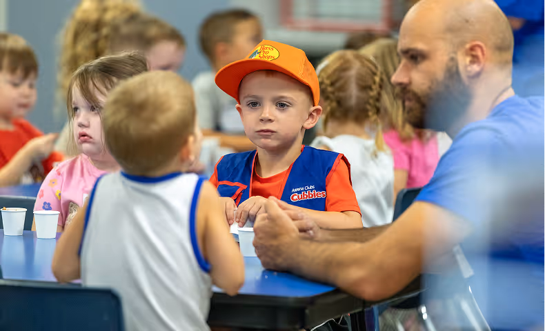 A group of children and a man sitting around a table with drinks in small cups, with one child wearing an orange hat and a vest labeled Cubbies.