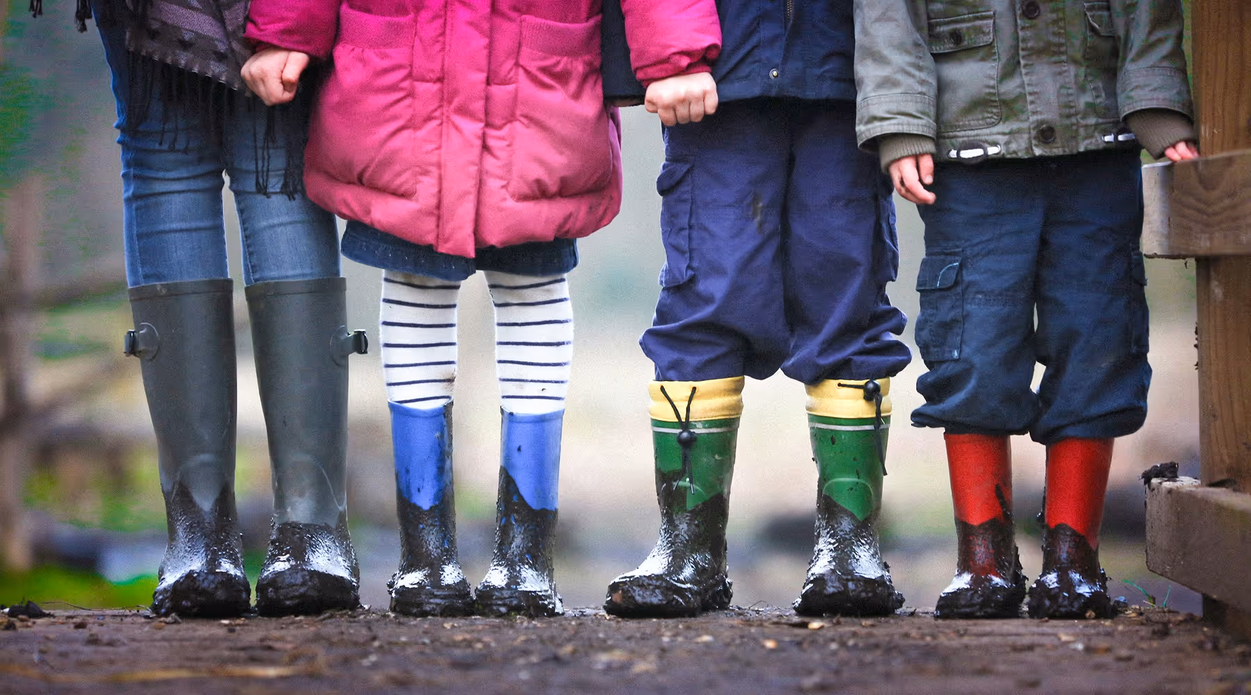 Four children wearing colorful muddy rain boots standing side by side outdoors.
