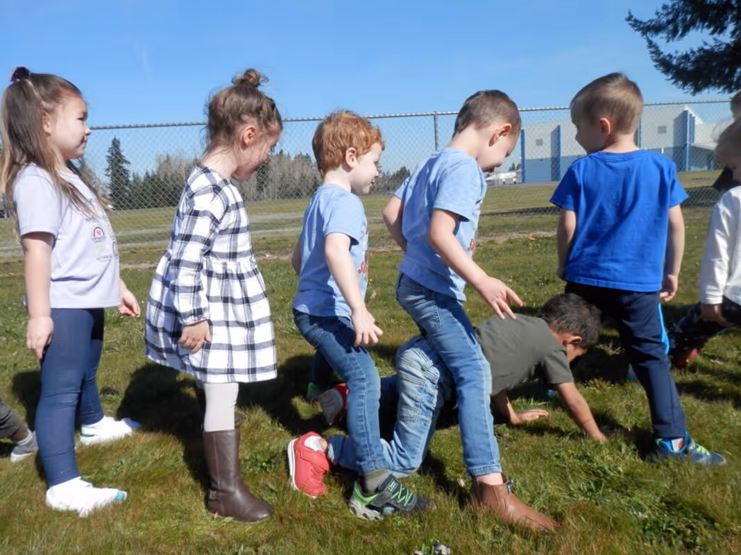 Group of young children playing and walking on grass outside on a sunny day near a chain-link fence.