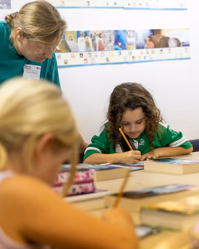 A volunteer assists a young curly-haired child writing with a pencil at a table with books while another child works in the foreground.