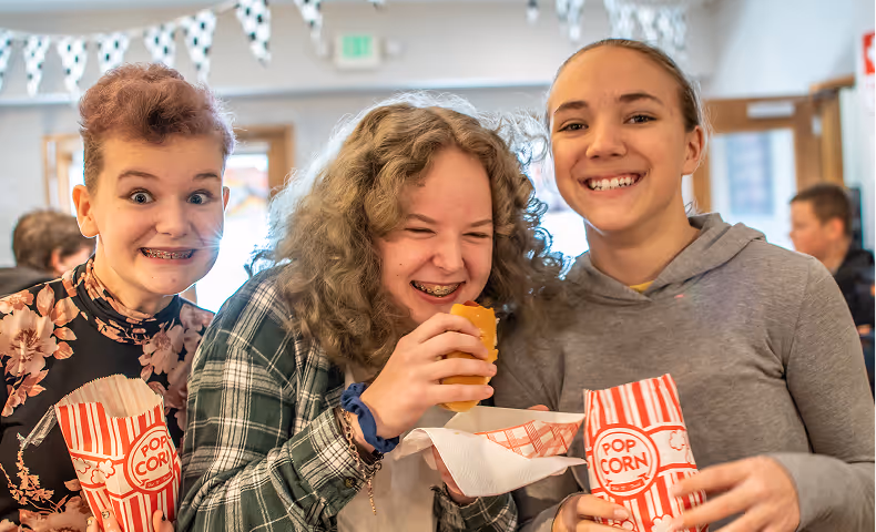 Three smiling children with braces holding popcorn and a hot dog in a festive indoor setting.