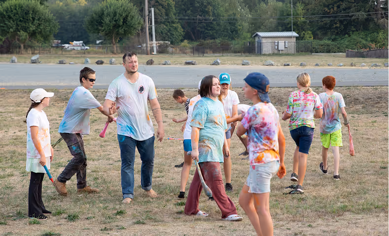 Group of children and an adult playing baseball outside on dry grass, wearing colorful tie-dye shirts.