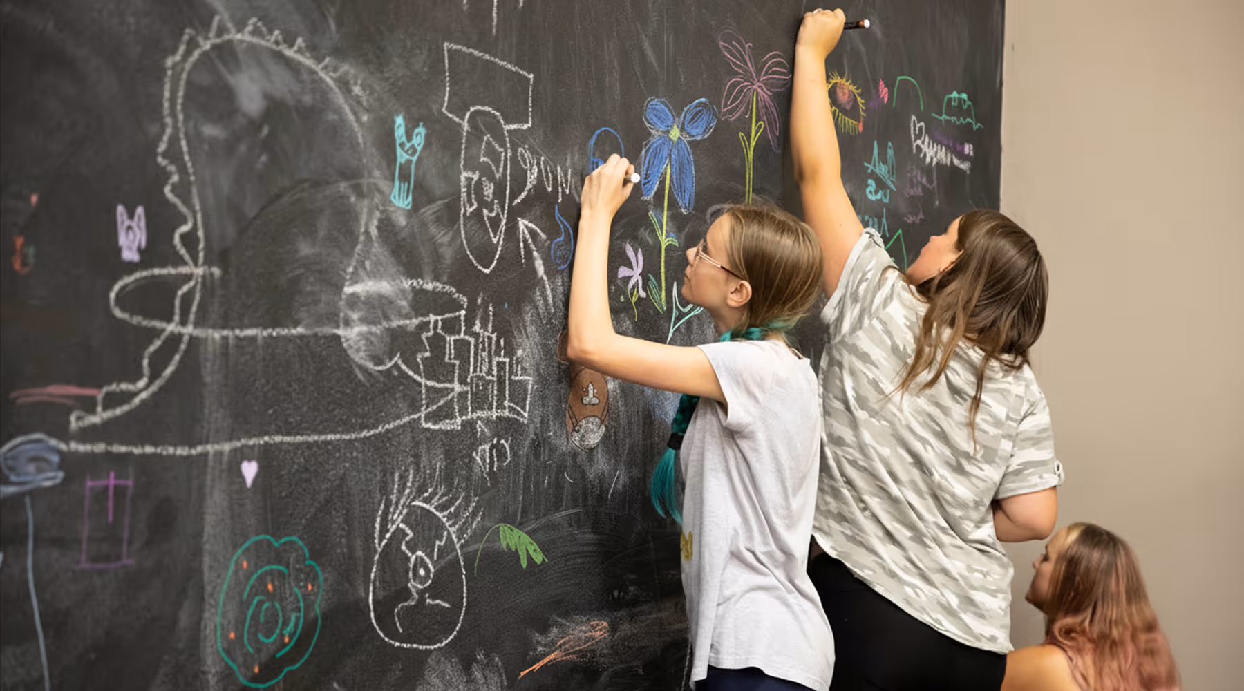 Three children drawing colorful pictures like flowers and faces on a large black chalkboard wall.
