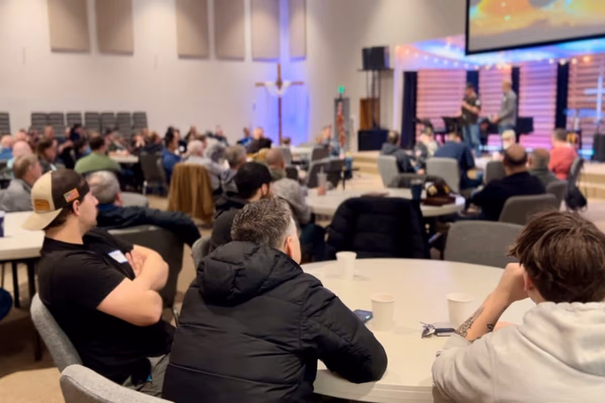 People seated at round tables attentively listening to speakers on stage.