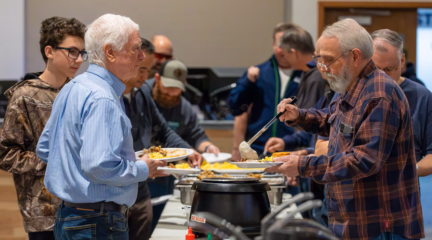 A group of men in casual clothing serving themselves food from a buffet table indoors.