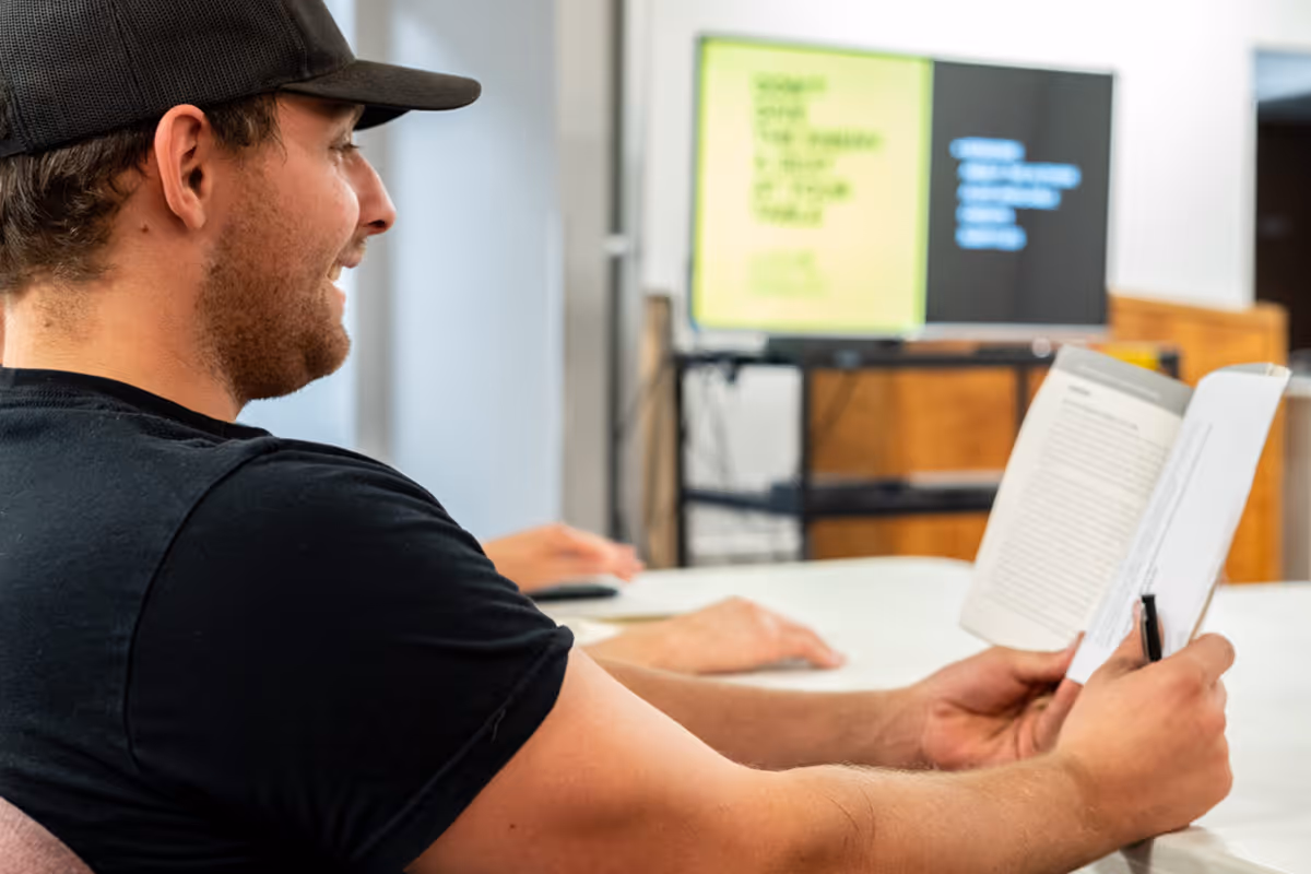 Man in a black shirt and cap reading a bible while sitting at a table with a blurred screen in the background.