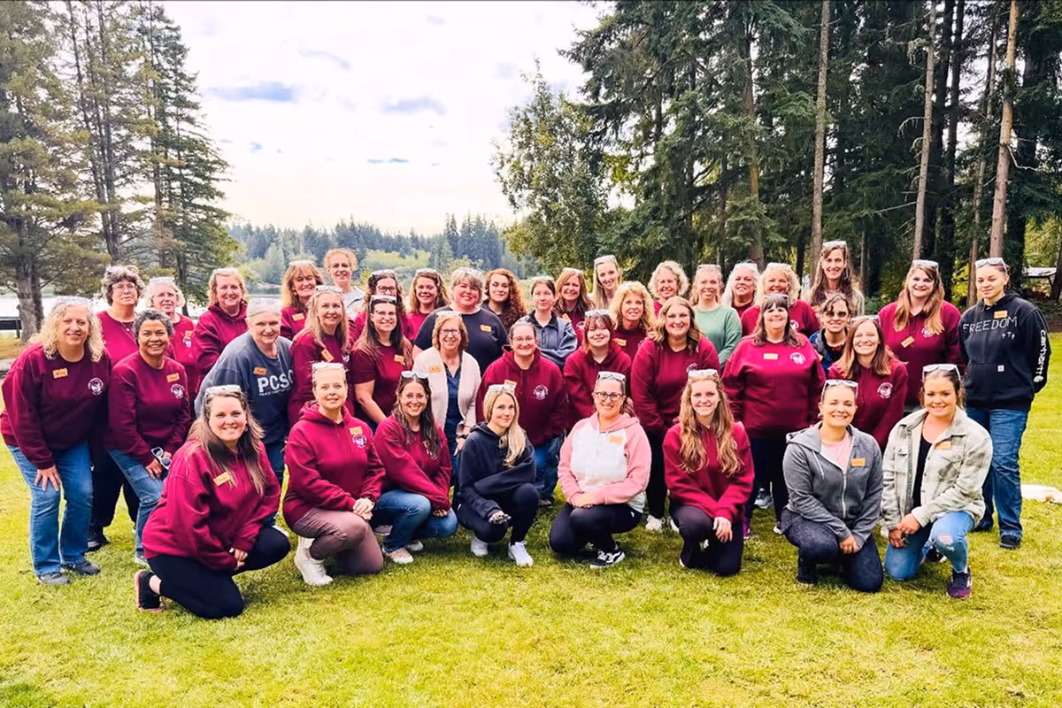 Large group of women gathered outdoors on green grass, many wearing matching maroon sweatshirts, with trees and a lake in the background.