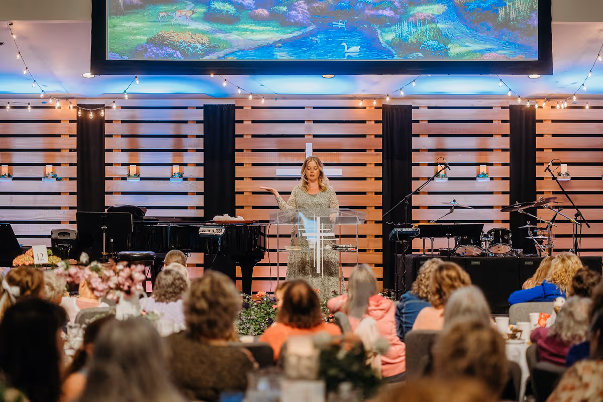 Woman speaking at a podium with a cross symbol in a church with an audience seated at tables and musical instruments in the background.