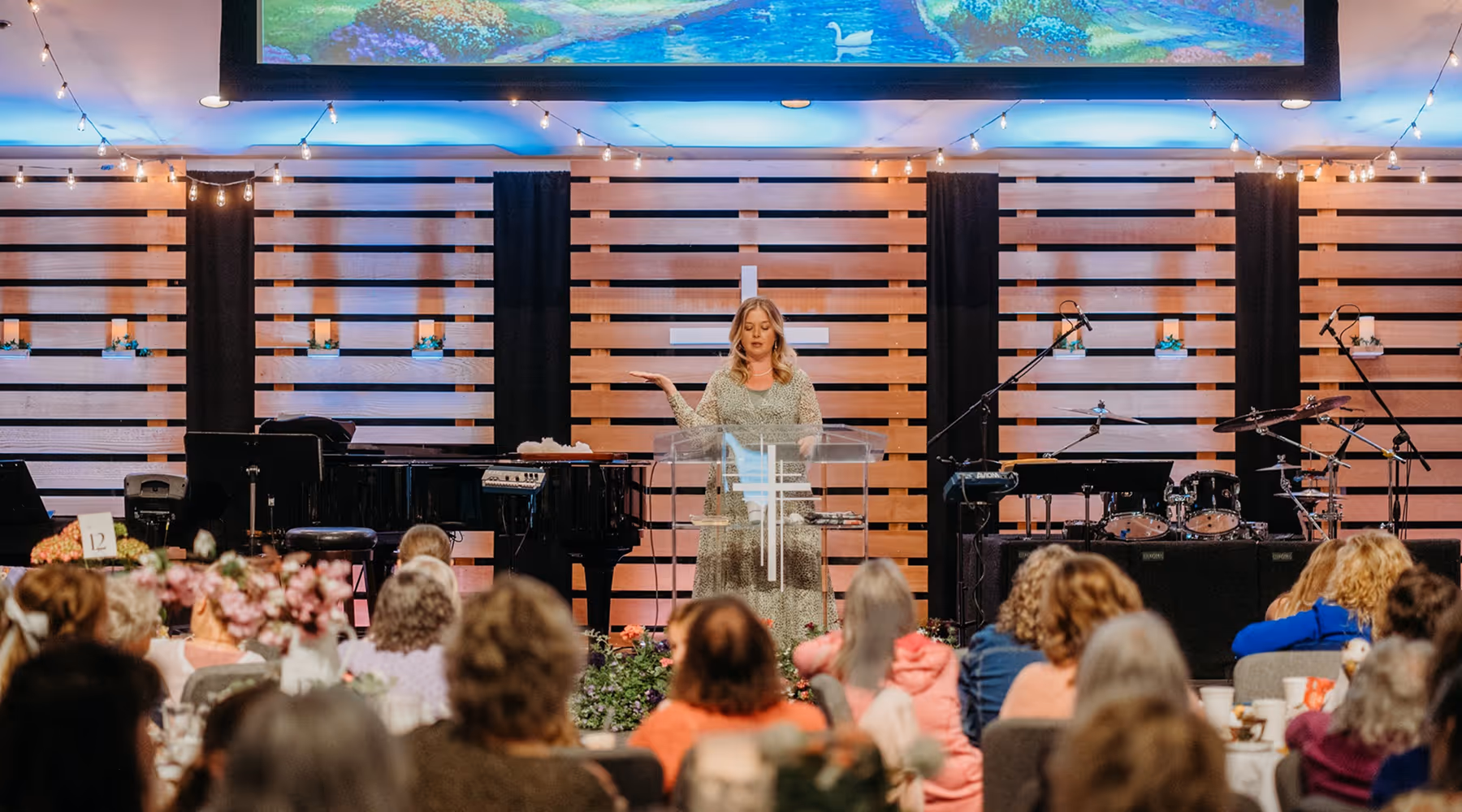 Woman speaking behind a clear podium in a church setting with a wooden slat wall, cross, piano, and drum set, addressing an audience.