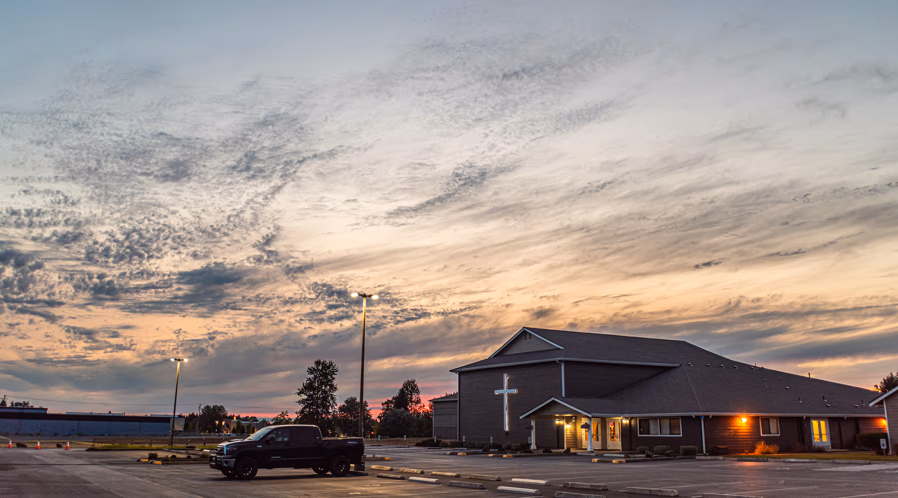 Church building with illuminated cross and a black pickup truck in an empty parking lot at sunset with cloudy sky.