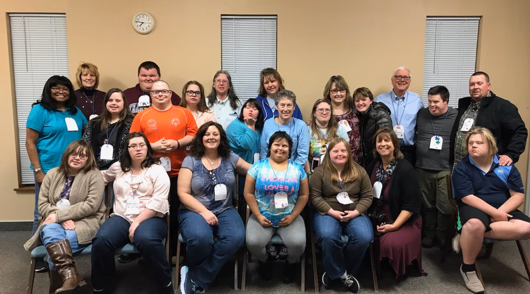 Group photo of diverse adults, some with disabilities, smiling indoors against beige walls with blinds and a clock.
