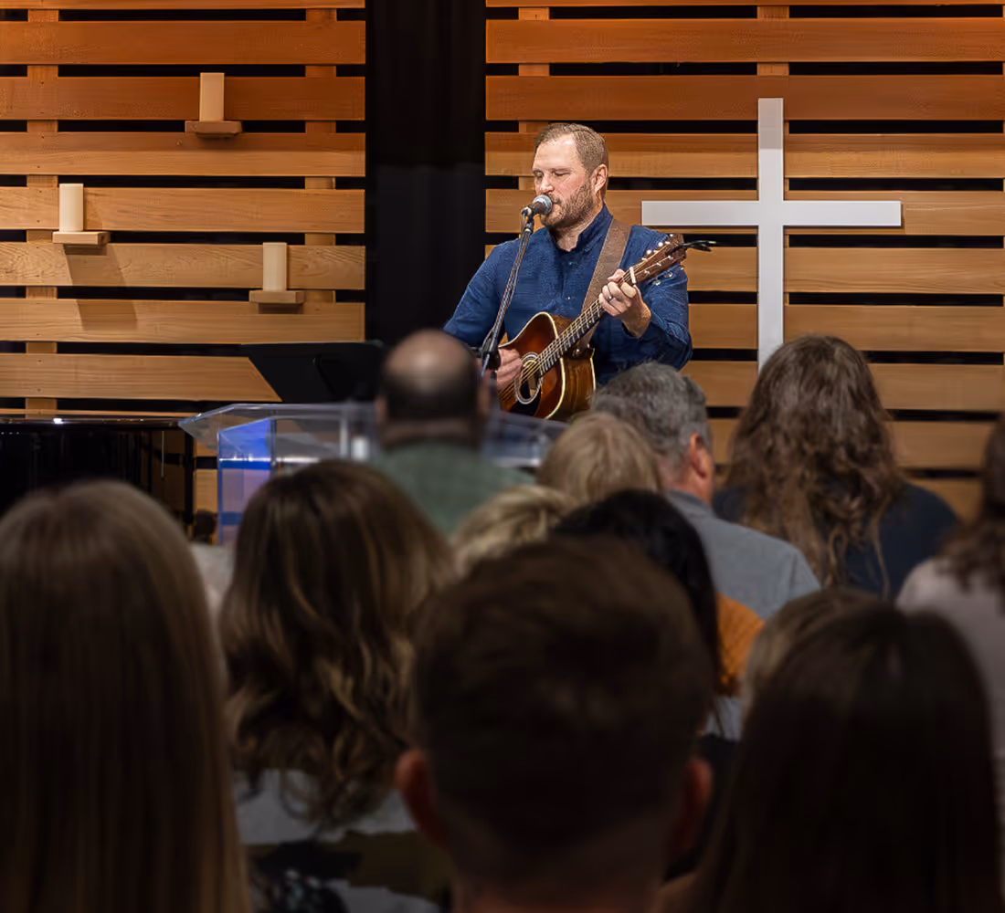 Man singing and playing acoustic guitar on stage in front of an audience inside a church with a white cross on a wooden wall.