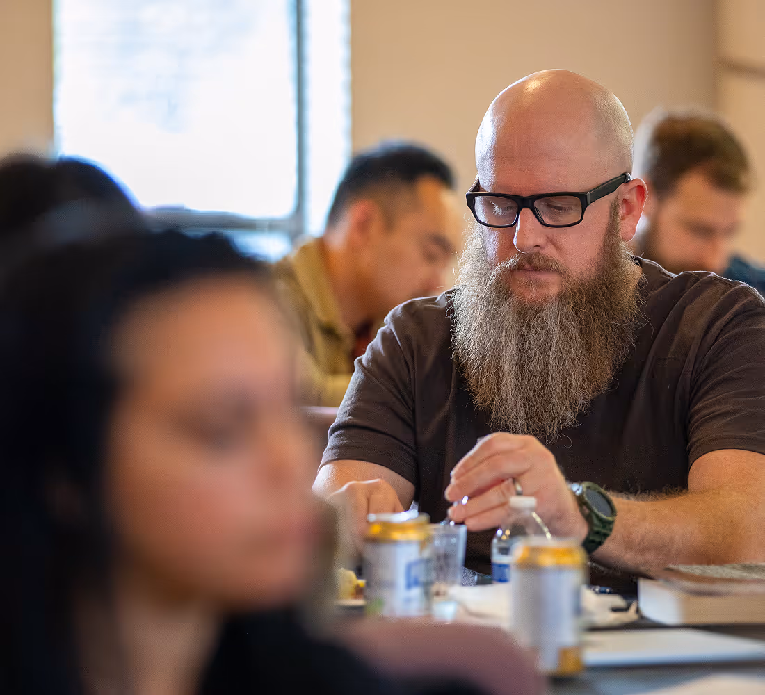 Bald man with glasses and long beard sitting at a table with drinks and food, focused on an activity indoors.