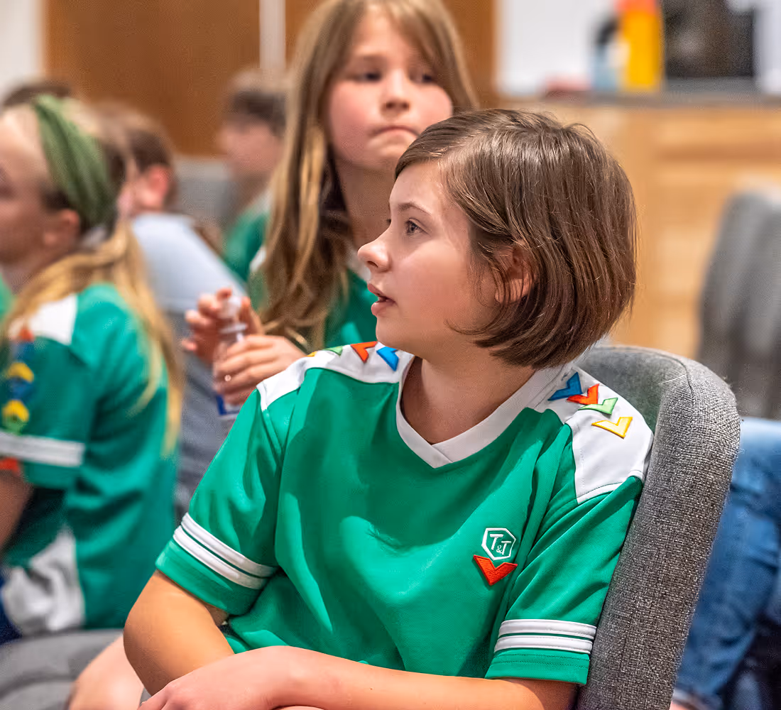 Group of children in green shirts sitting indoors, focused on something off-camera.