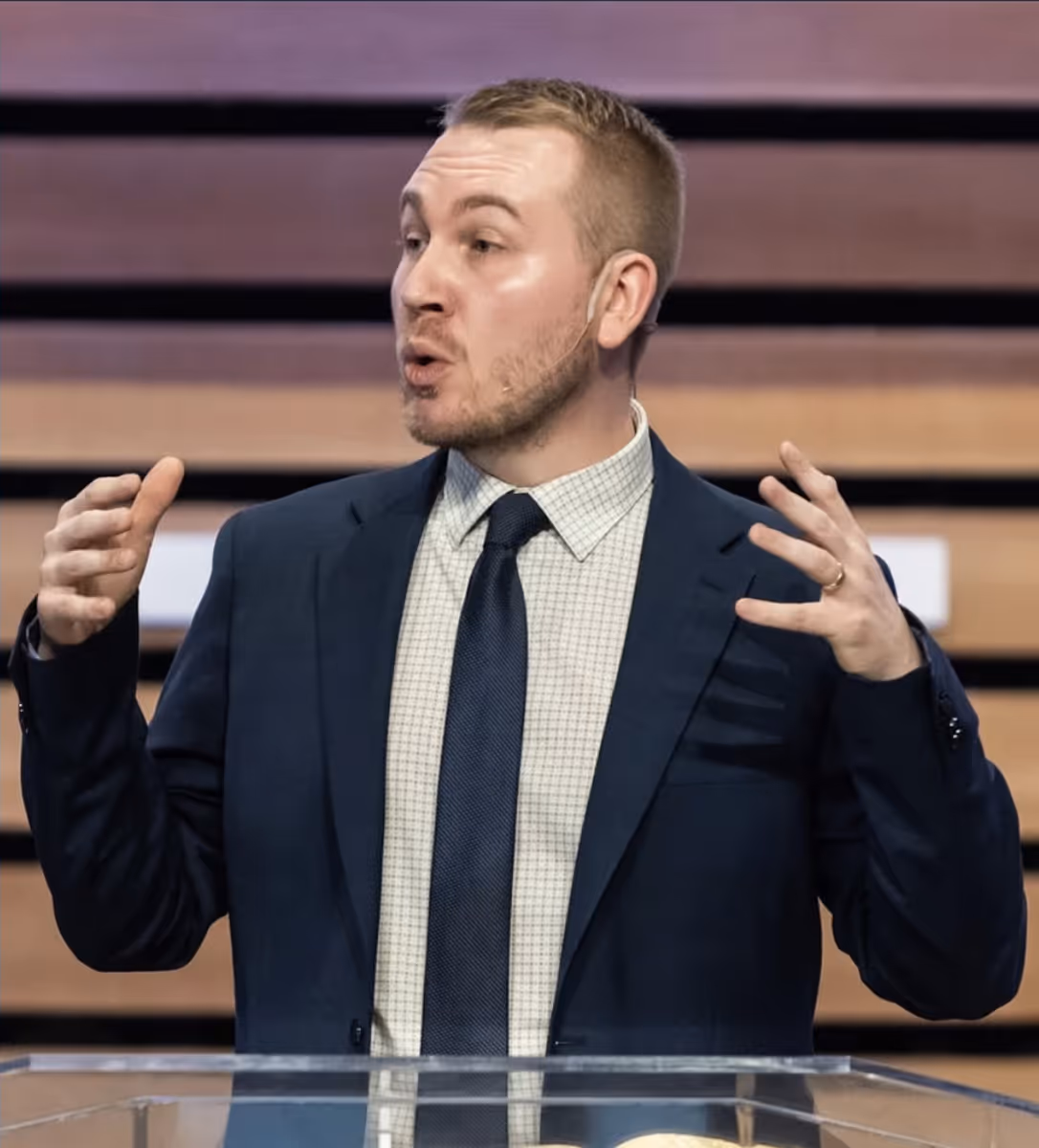Man in a suit speaking and gesturing with hands behind a clear podium.