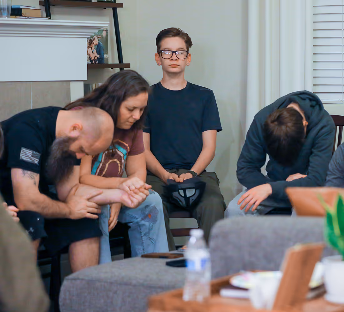 A group of people seated indoors, heads bowed and hands clasped in a moment of prayer or reflection.