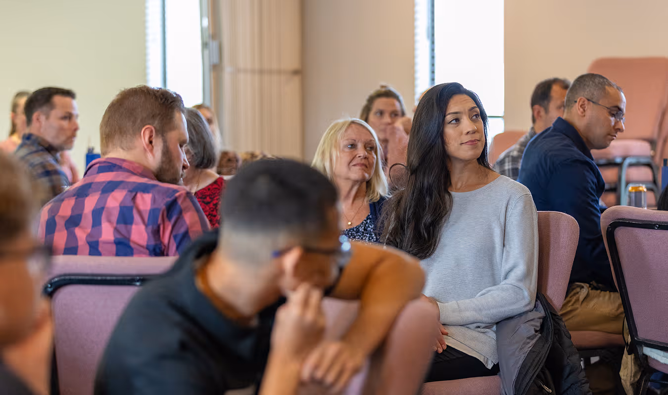 Group of diverse adults seated in a room, attentively listening, with a woman in a gray sweater in focus.