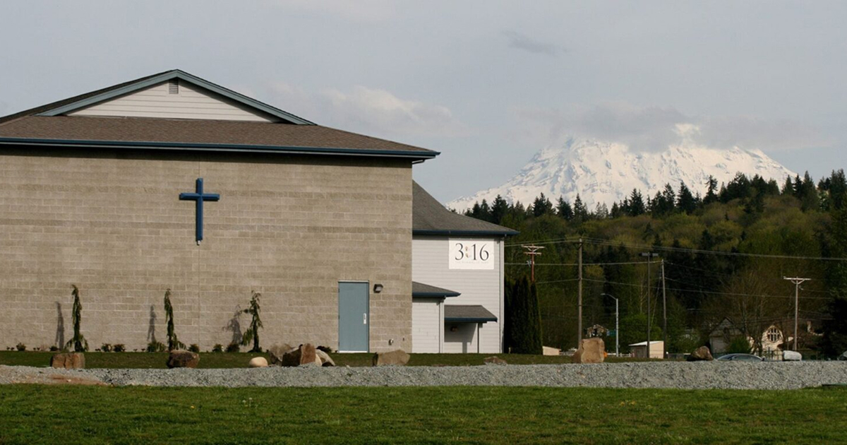 Side view of a church building with a blue cross, trees, and a snow-capped mountain in the background.