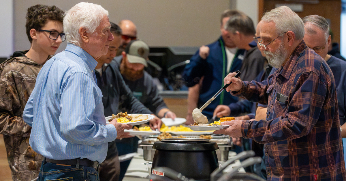 Men in line serving themselves food at a buffet with dishes like corn, vegetables, and biscuits.