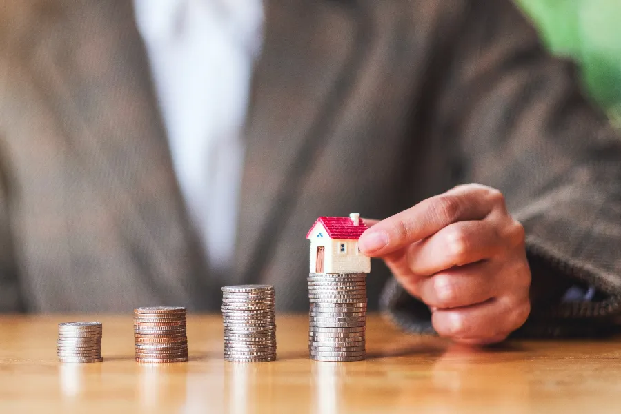 Hand placing a model house on a growing stack of coins.