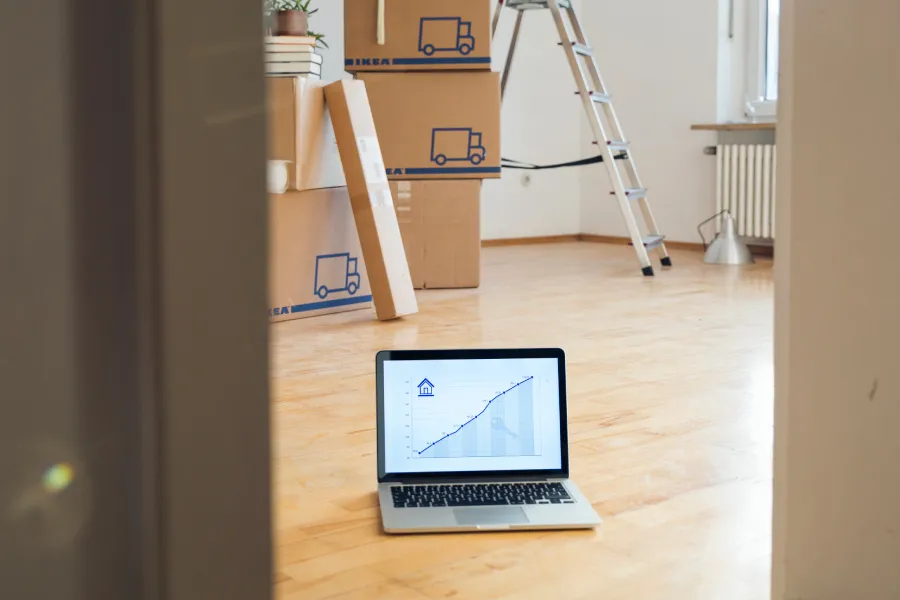 A laptop sitting on a wooden floor displaying an upward line graph, with moving boxes and a stepladder in the background.