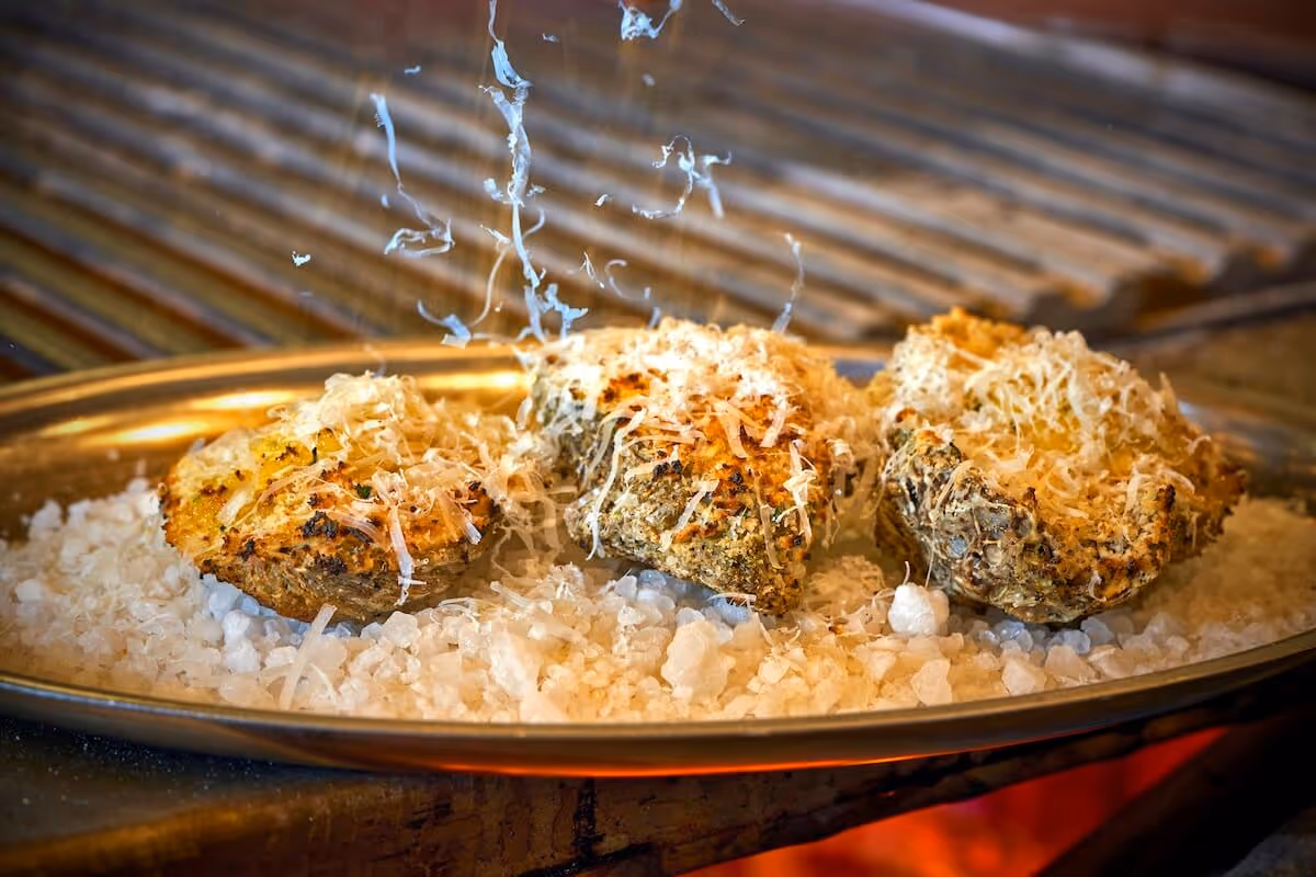 Three grilled oysters topped with melted cheese on a metal platter over coarse salt.