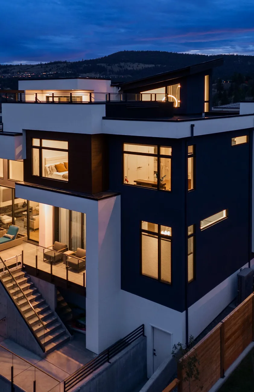Modern two-story house at dusk with illuminated windows, outdoor seating area, and staircase leading down.