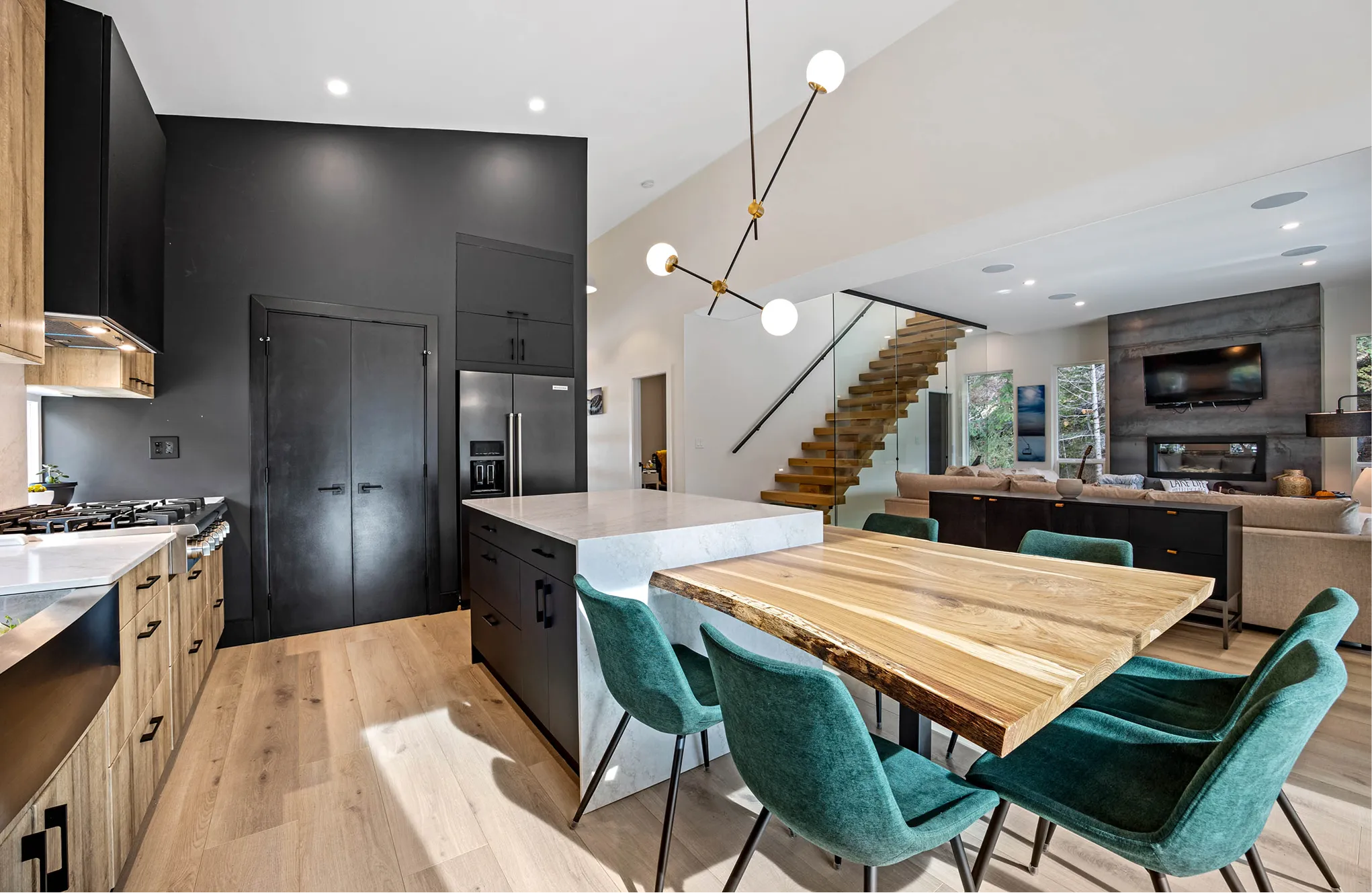 Modern open kitchen and living room with black and wood cabinetry, a marble island, wooden dining table with teal chairs, and floating wooden stairs with glass railing.