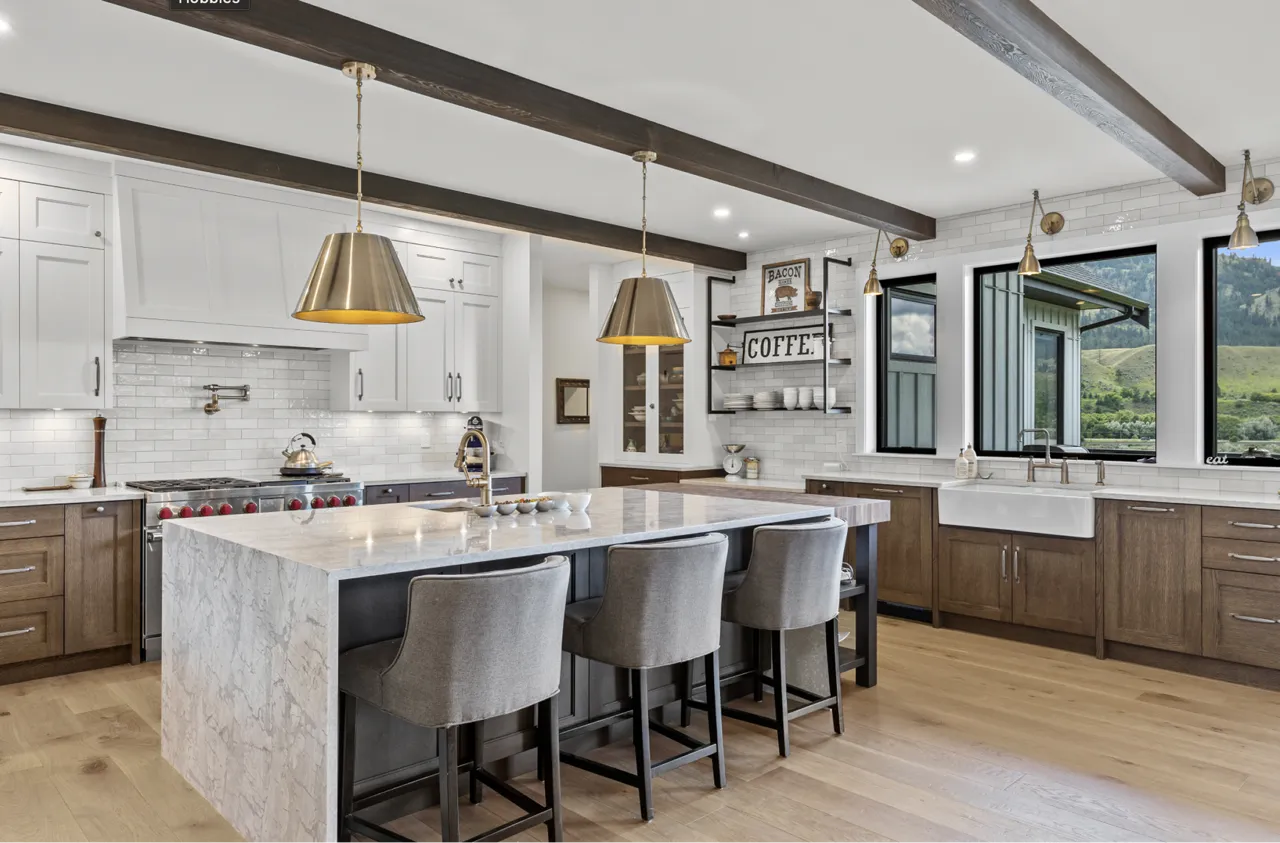 Modern kitchen with a large marble island, three gray upholstered chairs, white upper cabinets, wooden lower cabinets, and pendant lights hanging from a ceiling with dark wooden beams.