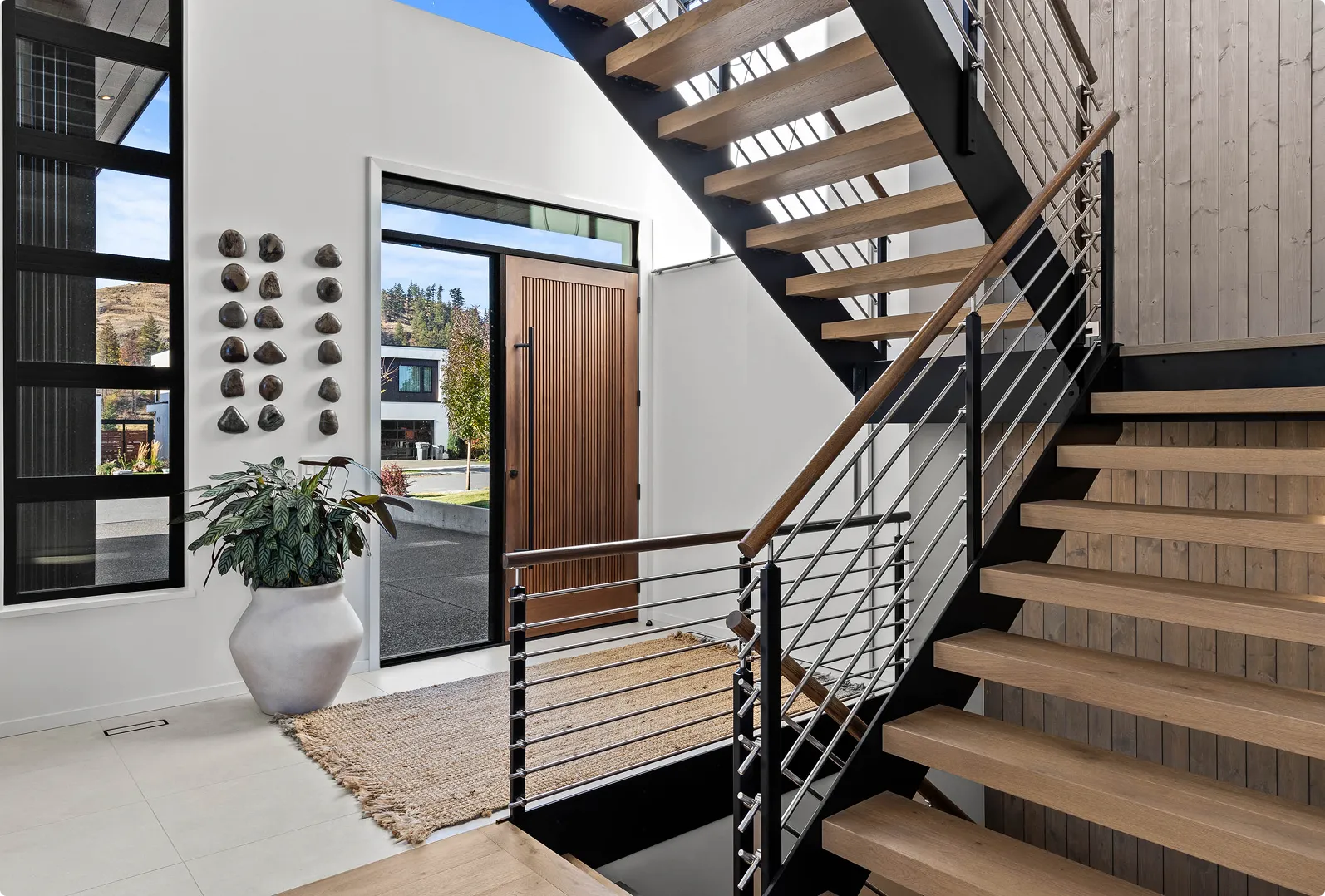 Modern interior entryway with wooden front door, large plant in white pot, and staircase with metal railing and wooden steps.