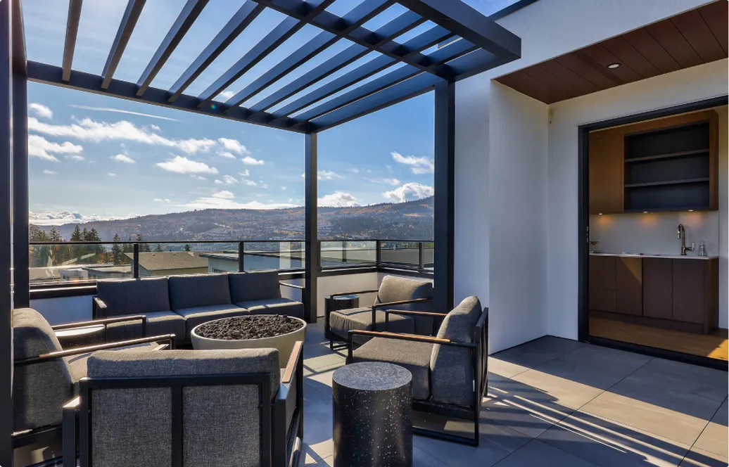 Modern rooftop patio with gray cushioned chairs, a central fire pit, and a pergola overlooking a landscape with hills under a blue sky.