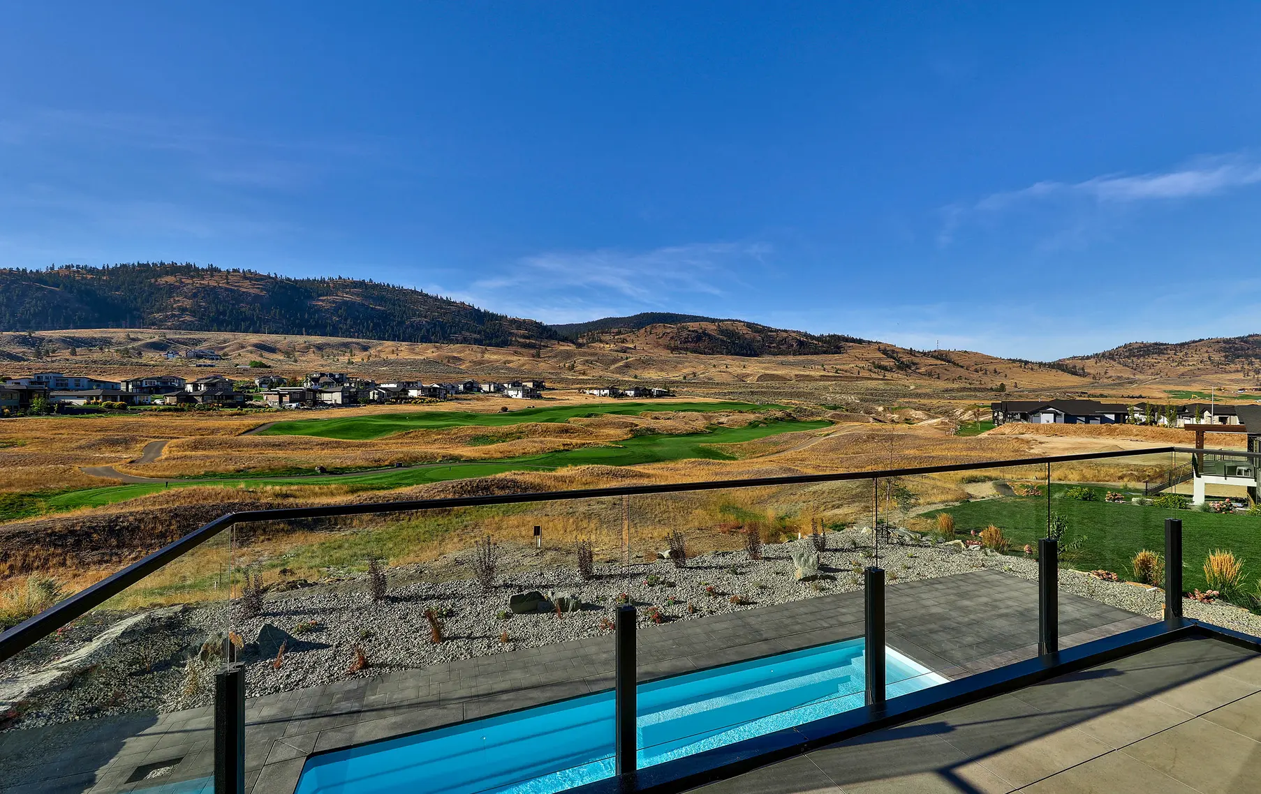 View from a balcony with glass railing overlooking a swimming pool, landscaped yard, golf course, and hills under a clear blue sky.