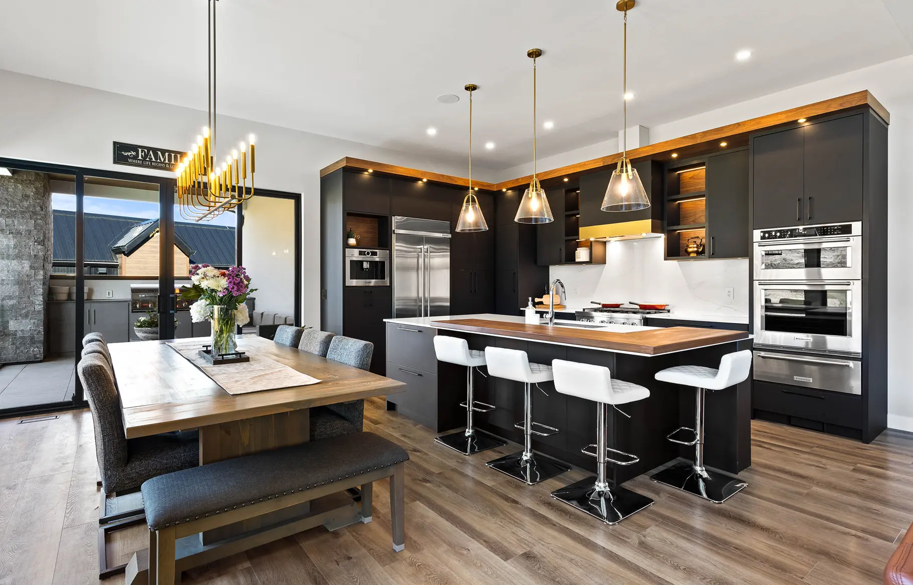 Modern kitchen and dining area with dark cabinetry, wood countertops, white bar stools, and a wooden dining table with gray chairs and a flower vase.