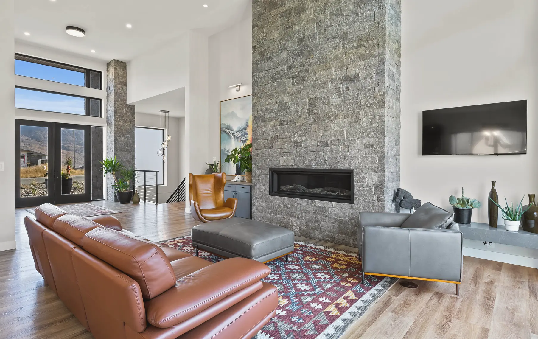 Modern living room with a large stone fireplace, brown leather sofa, gray armchair, orange accent chair, and a patterned area rug.